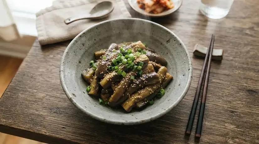 Overhead view of steaming Gaji-muchim in grey ceramic bowl with glossy sesame oil, sesame seeds, and green onion garnish